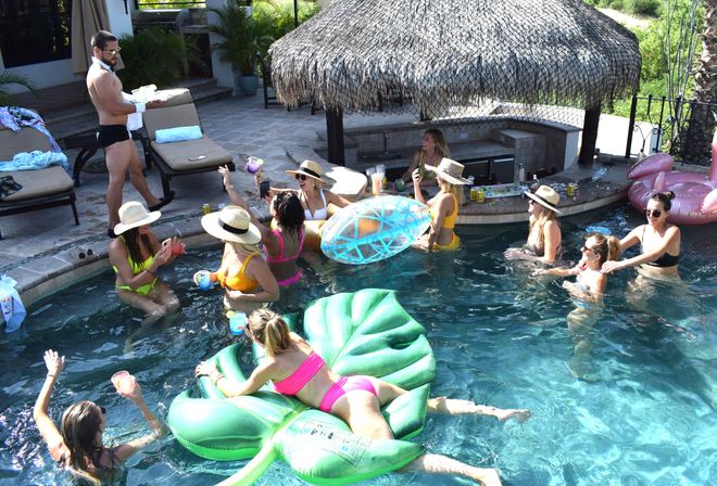 Sunny backyard pool party with a group of friends in colorful bikinis and sun hats, people enjoying drinks on a green leaf float and clear inflatable near a thatched cabana and poolside lounge chairs.