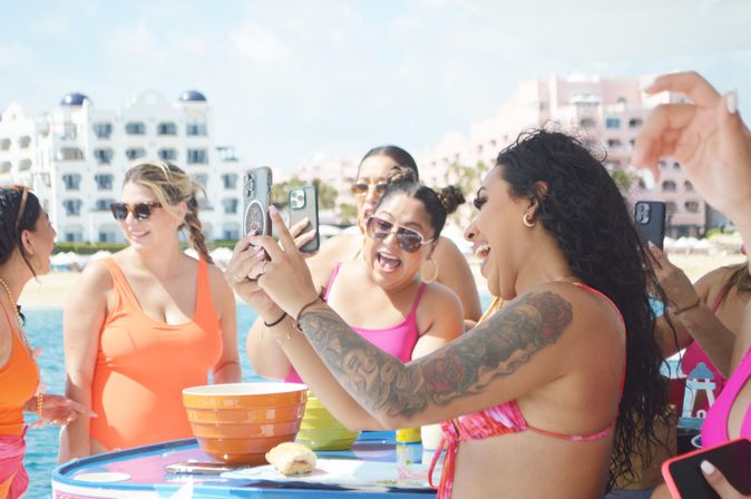 Laughing group of women in colorful swimsuits taking selfies at a sunny beachfront resort, gathered around a table with bowls and snacks with ocean and hotel buildings in the background.