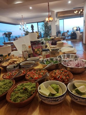 Ocean-view indoor spread of Mexican-style toppings ready for tacos: guacamole, pico de gallo, lime wedges, sliced red onion and tortilla chips on a wooden counter at sunset.