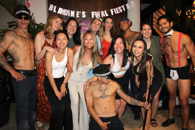 Smiling group of friends posing under a 'final fiesta' banner at an indoor celebration, with two shirtless male performers in police hats and playful costumes.