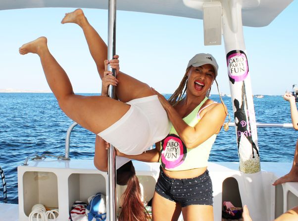 Two women laughing on a sunny boat party in Cabo — one hanging upside down on a dance pole while a friend steadies her against a bright blue ocean backdrop.