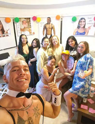 Indoor living-room party group selfie: smiling women gathered with a shirtless male entertainer in bow tie and cuffs, colorful balloons, posters and drinks on a table.