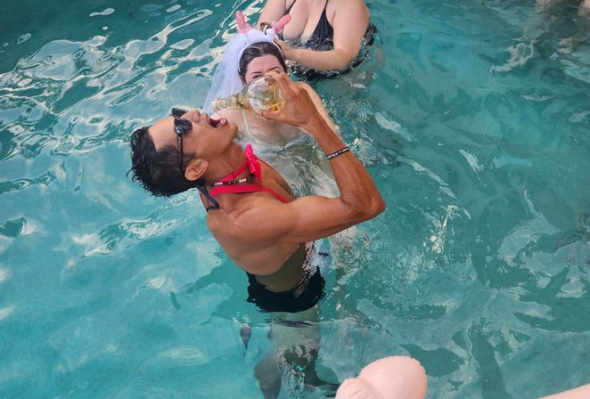 Fun pool party scene: a person in sunglasses and a red top tilts a bottle into their mouth while others, including someone wearing a white bridal-style veil headband, float in clear turquoise swimming pool water.