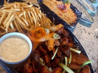 Platter of crispy fried chicken wings garnished with celery sticks and carrot ribbons, thin golden French fries with creamy ranch dip, and a side of seasoned rice on blue serving dishes on a stone countertop.