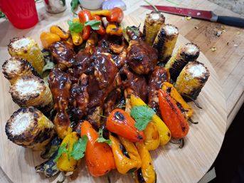 Mouthwatering grilled BBQ chicken drumsticks glazed in dark sauce on a round wooden board, surrounded by charred corn on the cob and blistered orange and yellow mini sweet peppers, garnished with cilantro — summer barbecue platter.