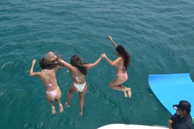 Three friends in bikinis holding hands as they leap off a boat into clear turquoise ocean, captured mid-air in a playful summer jump.