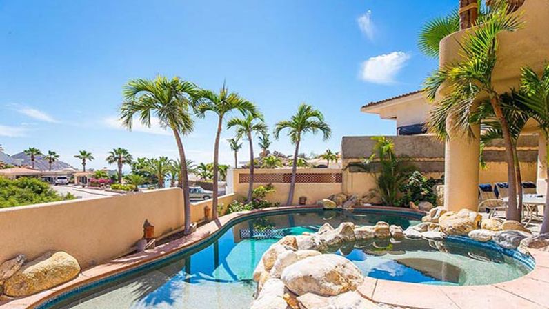 Sun-drenched beachside pool and spa with rock features, swaying palm trees, stucco patio and distant ocean view under a bright blue sky.