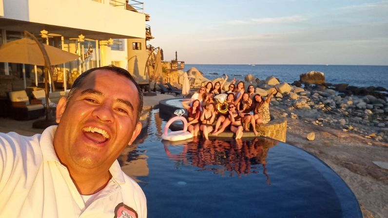 Man taking a joyful selfie in the foreground while a large group in swimsuits poses on the edge of an infinity pool with a pink pool float and brass instrument, beachfront rocky shoreline and ocean at sunset.