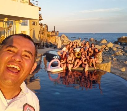 Man taking a joyful selfie in the foreground while a large group in swimsuits poses on the edge of an infinity pool with a pink pool float and brass instrument, beachfront rocky shoreline and ocean at sunset.