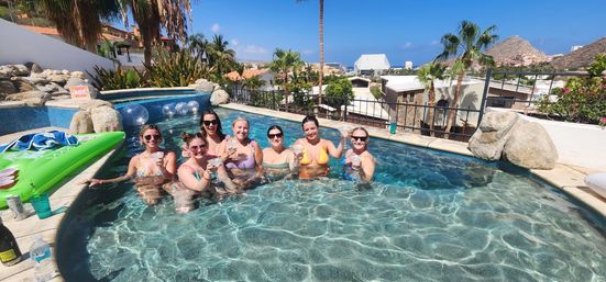 Group of friends in bikinis raising drinks in a sunny villa pool with palm trees and coastal hillside homes under a clear blue sky.