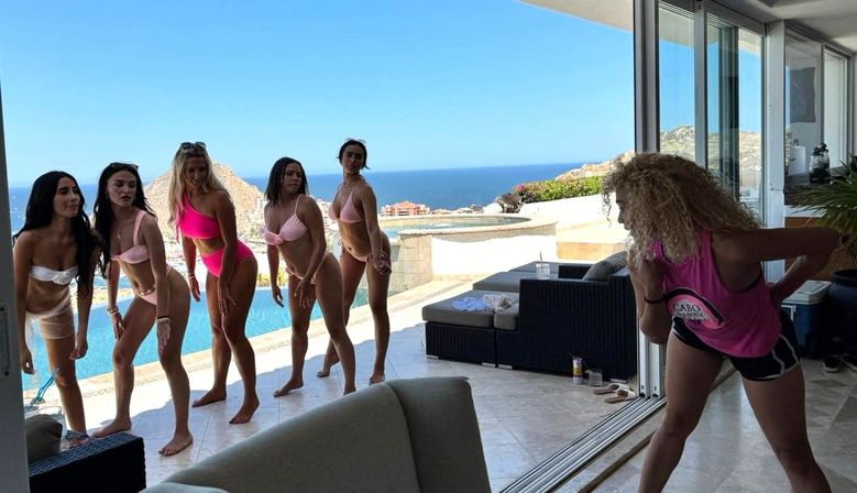 Six women in bikinis lined up by an infinity pool at a sunlit beachfront villa overlooking the ocean, while a photographer in a pink tank top directs their pose.