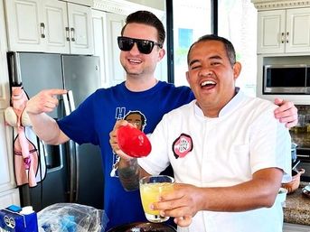 Two smiling men in a bright modern kitchen — one wearing a chef’s coat holding a red bell pepper and a glass of orange juice, the other in sunglasses and a blue T‑shirt pointing at him, stainless steel fridge and white cabinets behind them.