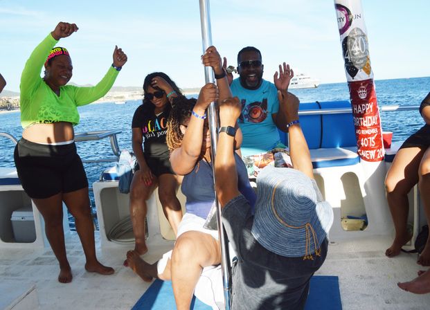 Group of adults enjoying a sunny boat party, dancing around a pole and celebrating a birthday with ocean and a distant yacht in the background.