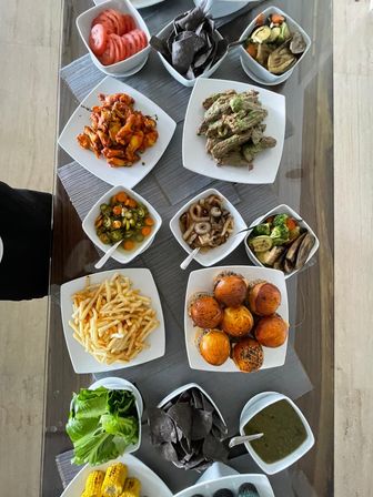 Overhead shot of a colorful home dining table spread with glossy mini burger sliders, shoestring fries, buffalo-style wings, grilled corn, blue corn chips, sliced tomatoes, pickled jalapeños, roasted vegetables and a green dipping sauce.