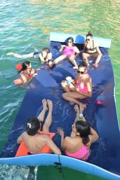 Six friends in colorful swimsuits lounging on a large blue floating mat in green water, sipping drinks and chatting on a sunny summer boat day