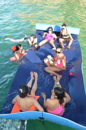 Six friends in colorful swimsuits lounging on a large blue floating mat in green water, sipping drinks and chatting on a sunny summer boat day