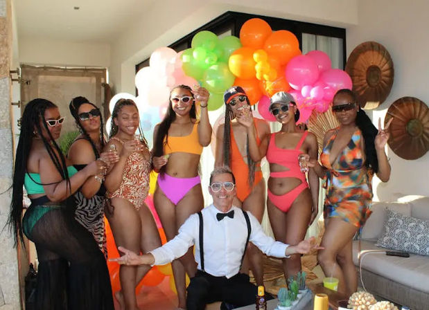 Colorful pool party on a sunny villa patio: group of friends in vibrant swimsuits and sunglasses with a man in suspenders and bow tie posing in front of a rainbow balloon garland, holding drinks.