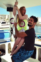 Red-haired woman laughing as she clings to a dance pole on a sunny party boat while a smiling man holds her legs, with yachts and a coastal marina in the background.