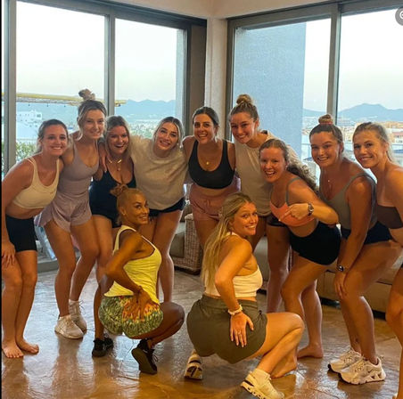 Smiling group of young women in casual activewear posing and squatting together in a sunlit apartment living room with floor-to-ceiling windows showing distant mountains and urban skyline.
