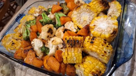 Colorful roasted vegetable medley in a glass baking dish: charred corn on the cob rounds, seasoned sweet potato cubes, broccoli and cauliflower florets — golden and slightly charred, ready to serve.