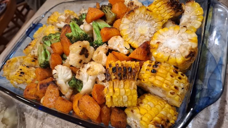 Colorful roasted vegetable medley in a glass baking dish: charred corn on the cob rounds, seasoned sweet potato cubes, broccoli and cauliflower florets — golden and slightly charred, ready to serve.