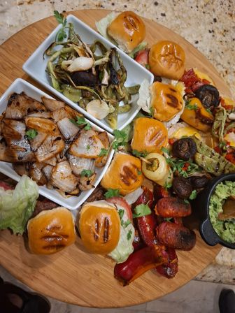 Round wooden appetizer board with mini sliders, charred peppers and sausages, roasted mushrooms and onions, grilled green vegetables, and a small bowl of guacamole