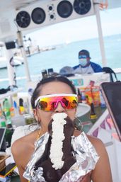 Person on a party boat biting a large chocolate novelty penis-shaped dessert topped with whipped cream, wearing mirrored sunglasses with a marina and turquoise ocean in the background.