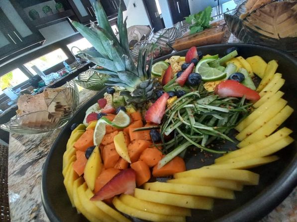 Vibrant tropical fruit platter on a kitchen island with a pineapple crown centerpiece surrounded by fanned mango slices, cubed papaya, cucumber ribbons, strawberries, blueberries and lime wedges.