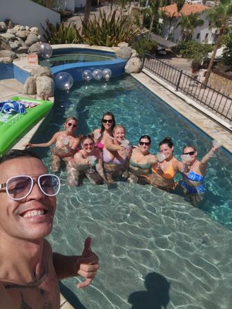 Sunny backyard pool party selfie — man giving thumbs-up in foreground, seven women in colorful swimsuits toasting cocktails in clear pool near a spa area and inflatable float.