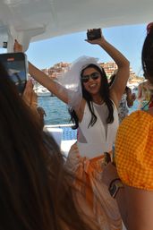 Bride-to-be wearing a veil and sunglasses, arms raised and dancing on a sunny boat party in a marina with waterfront buildings in the background