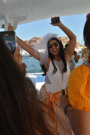 Bride-to-be in a white veil and sunglasses cheering on a sunlit party boat at a busy marina, holding a phone with friends in colorful summer outfits and waterfront buildings behind her.