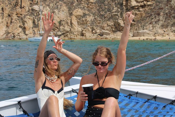 Two friends in swimsuits raising their arms and enjoying drinks on a boat hammock near a rocky coastal cove with turquoise water on a sunny vacation day
