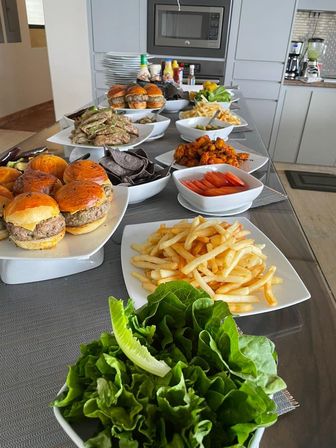Party-style spread on a modern kitchen island with slider burgers, French fries, fresh lettuce, blue corn chips, marinated meats, sliced tomatoes and assorted appetizers in white serving dishes.