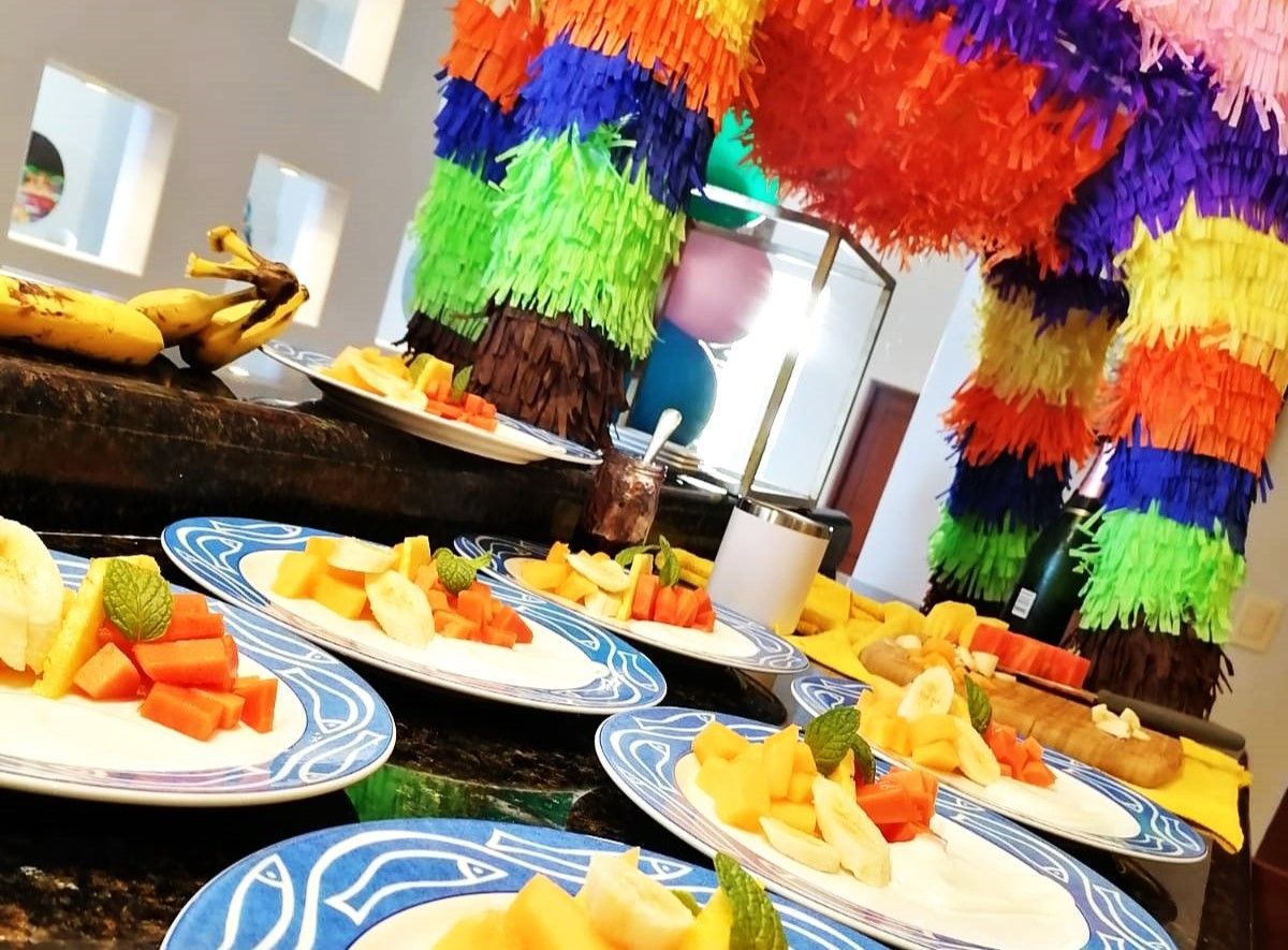 Party buffet counter with blue-rimmed plates of diced tropical fruit and mint garnish beneath a colorful rainbow piñata and balloons
