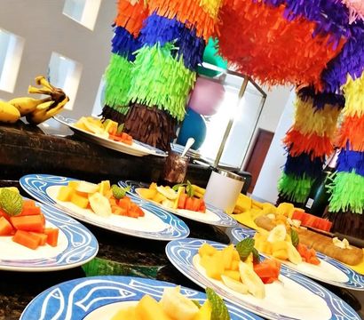 Party buffet counter with blue-rimmed plates of diced tropical fruit and mint garnish beneath a colorful rainbow piñata and balloons
