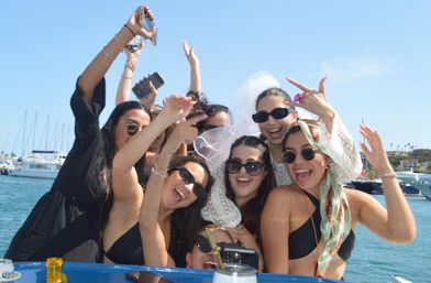 Laughing group of friends on a boat at a sunny marina celebrating a bachelorette party—sunglasses, veil, phones and yachts in the background.