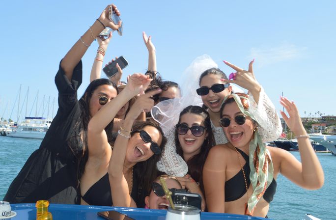 Laughing group of friends on a boat at a sunny marina celebrating a bachelorette party—sunglasses, veil, phones and yachts in the background.
