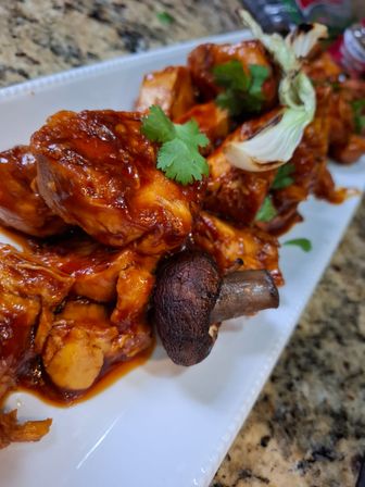 Close-up of sticky BBQ chicken skewers with grilled mushrooms and charred onion, garnished with cilantro on a white platter over a granite countertop
