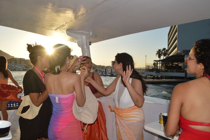 Group of women chatting and sipping drinks on a sunset boat near a busy marina, wearing colorful swimsuits and cover-ups with sun flare over the water.