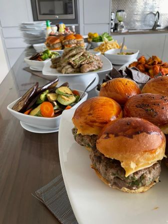 Mini cheeseburger sliders on glossy brioche buns in the foreground, with grilled vegetables, sliced steak, fries, wings and chips arranged as a crowd-pleasing buffet on a modern kitchen island.