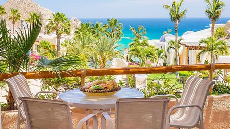 Sunlit beachfront balcony at a tropical resort: round table with fruit bowl and four chairs, lush palm gardens and turquoise ocean with rocky headland in the distance.