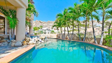 Turquoise curved backyard pool at a palm-lined villa patio with rock landscaping and desert hillside homes under a bright blue sky