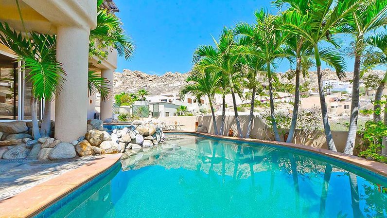 Turquoise curved backyard pool at a palm-lined villa patio with rock landscaping and desert hillside homes under a bright blue sky