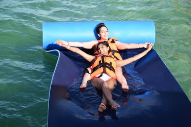 Two smiling women in orange life jackets lounging back-to-back on a blue floating mat in green water, arms outstretched enjoying a sunny water activity.