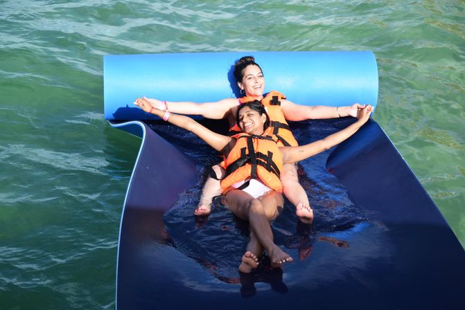 Two smiling women in orange life jackets lounging back-to-back on a blue floating mat in green water, arms outstretched enjoying a sunny water activity.