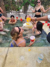 Outdoor backyard hot tub party with friends in swimsuits, someone pouring a pitcher of citrus cocktail into a friend’s mouth as others laugh, red solo cups and greenery around the spa