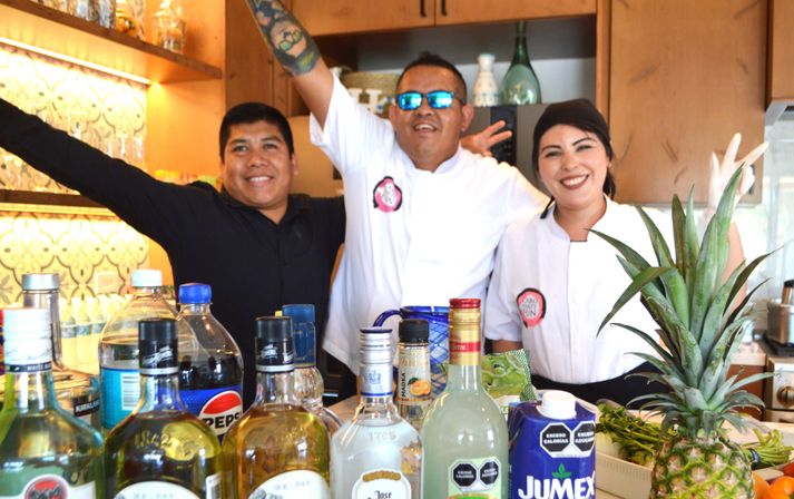 Three cheerful hospitality staff posing behind a restaurant bar and cocktail station with assorted liquor and mixer bottles, glassware, herbs and a pineapple garnish.