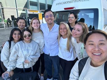 Smiling group of travelers in matching Cabo San Lucas shirts posing at an airport curb in front of a white shuttle van, ready for a group trip