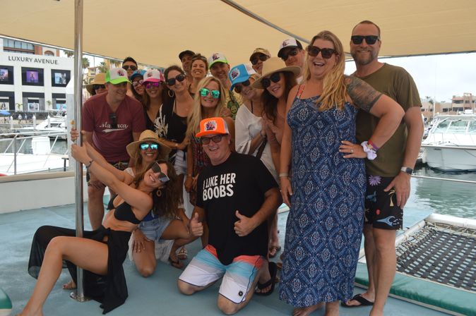 Smiling group of adults on a sun‑shaded catamaran at a marina, wearing hats, sunglasses and beachwear, posing for a fun vacation photo