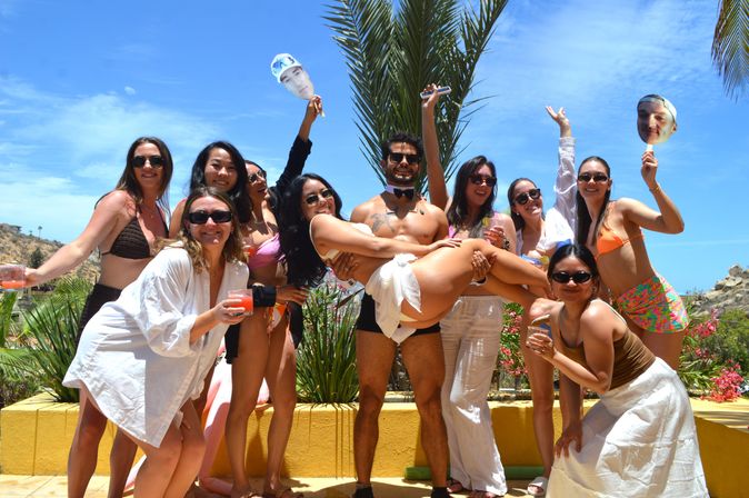 Group of friends at a sunny tropical poolside party with palm trees and blue sky, wearing swimwear and sunglasses, holding cocktails and face cutouts while a bow-tie–clad man lifts a woman in a white bikini.